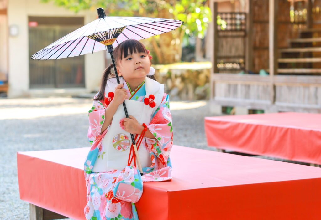 高鴨神社に七五三参りをしたピンク系の和装をした女の子が和傘を持って赤い椅子に座っている