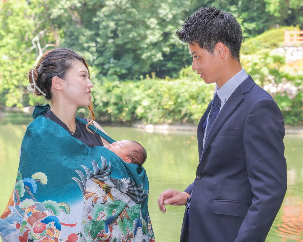 新緑の中の高鴨神社へお宮参り。緑の産着を纏い男の子を抱いた夫婦がお互いを見つめて微笑む