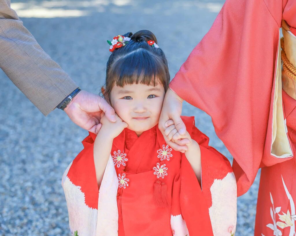 七五三参り　赤い和装の３歳女の子が両親に手を繋がれ顔に当てている様子