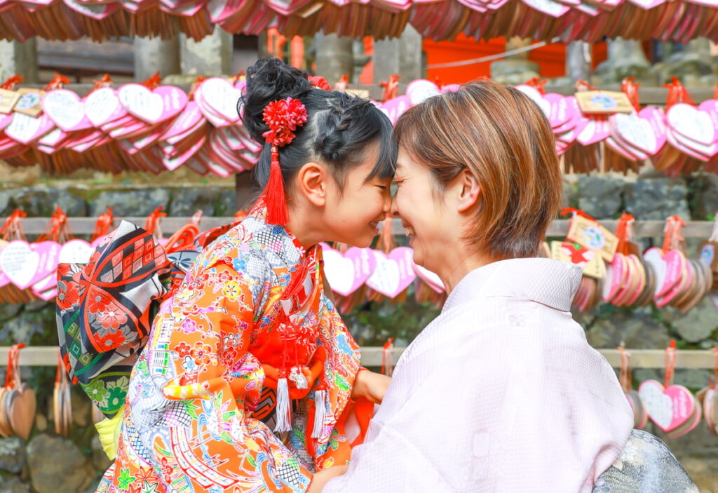 春日大社の夫婦大国社のハート型の絵馬の前で、七五三参りのオレンジの和装の女の子と、ピンクの和装をした母親がお互いのおでこをつけて楽しそうに笑っている様子