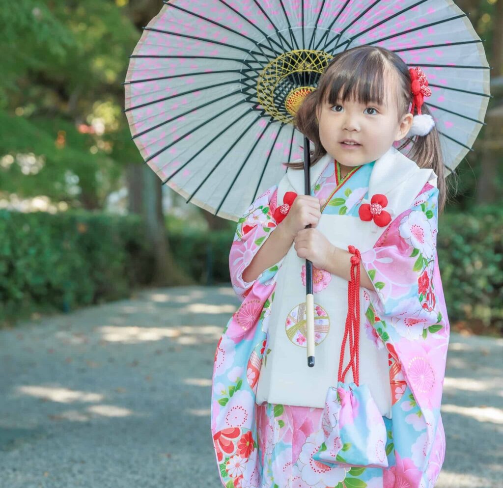 高鴨神社の真っ赤に紅葉したもみじの前で、七五三参りを終えた和装の女の子が笑顔でこちらを見ている