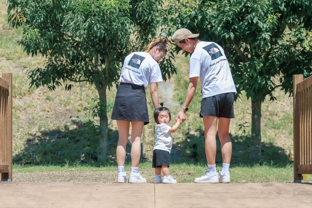 公園で白いTシャツに半パンのお揃いコーデの親子が子どもと手を繋いで微笑む様。真ん中の一歳の男の子はこちらへ振り返っている。